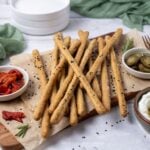 A close up photo of sourdough grissini garnished with black and white sesame seeds. The crunchy sourdough breadsticks are piled on a board surrounded by some antipasto type food.