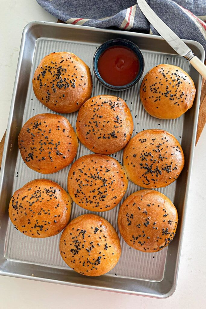 A silver baking tray containing 9 sourdough discard hamburger buns topped with black sesame seeds. 