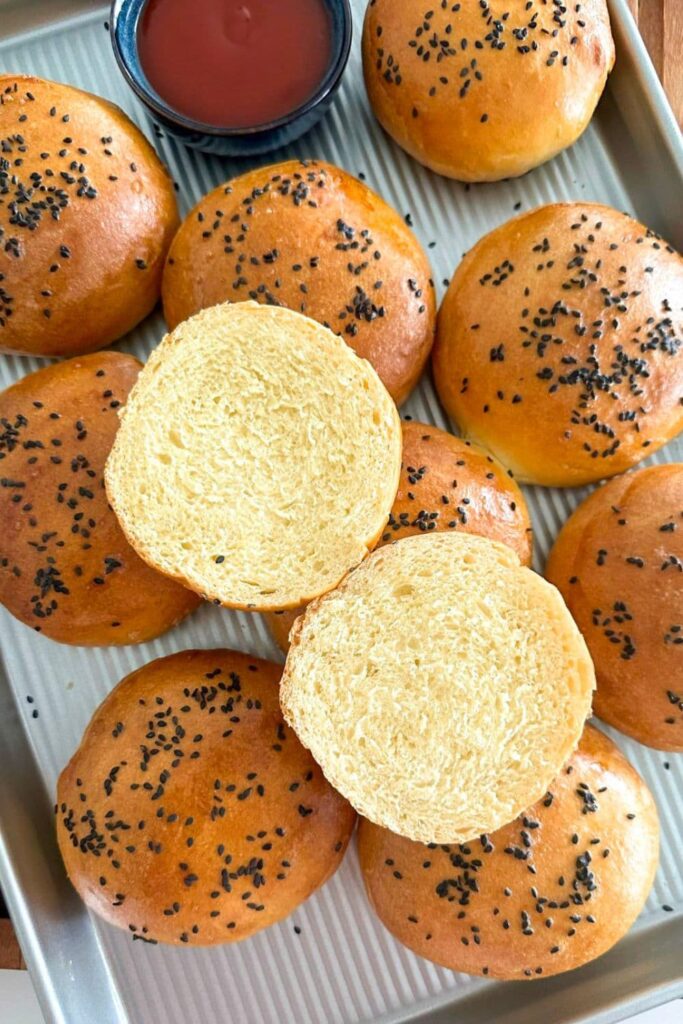 A silver baking tray holding sourdough discard burger buns coated in black sesame seeds. One of the sourdough discard hamburger buns has been sliced open and placed on top so you can see the soft crumb inside.