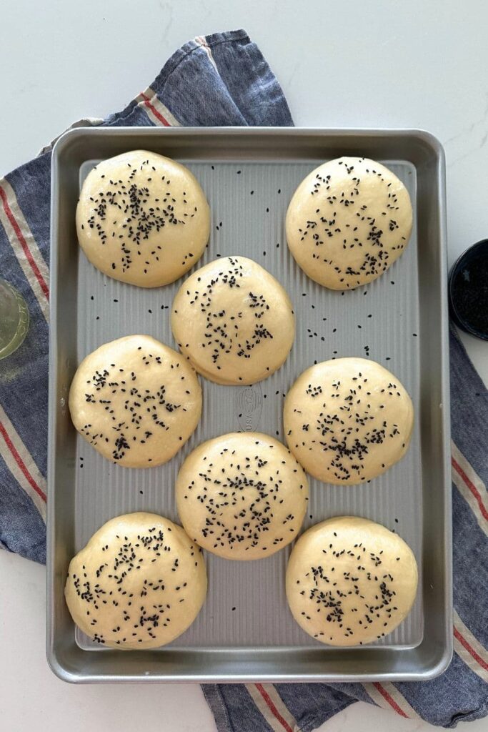 A silver baking tray containing 9 unbaked sourdough discard hamburger buns topped with black sesame seeds.