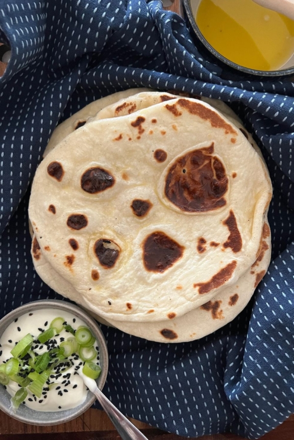 A stack of sourdough naan sitting on a dark blue dish towel. On the bottom left of the photo there is a small bowl of yogurt garnished with spring onions.