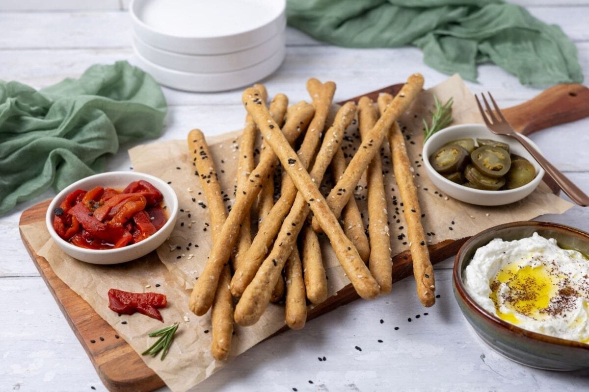 Horizontal image of crunchy sourdough breadsticks displayed on a rectangle serving board. There are some roasted red peppers and jalapenos on the board, as well as some whipped feta dip to the right. The board is garnished with fresh rosemary.
