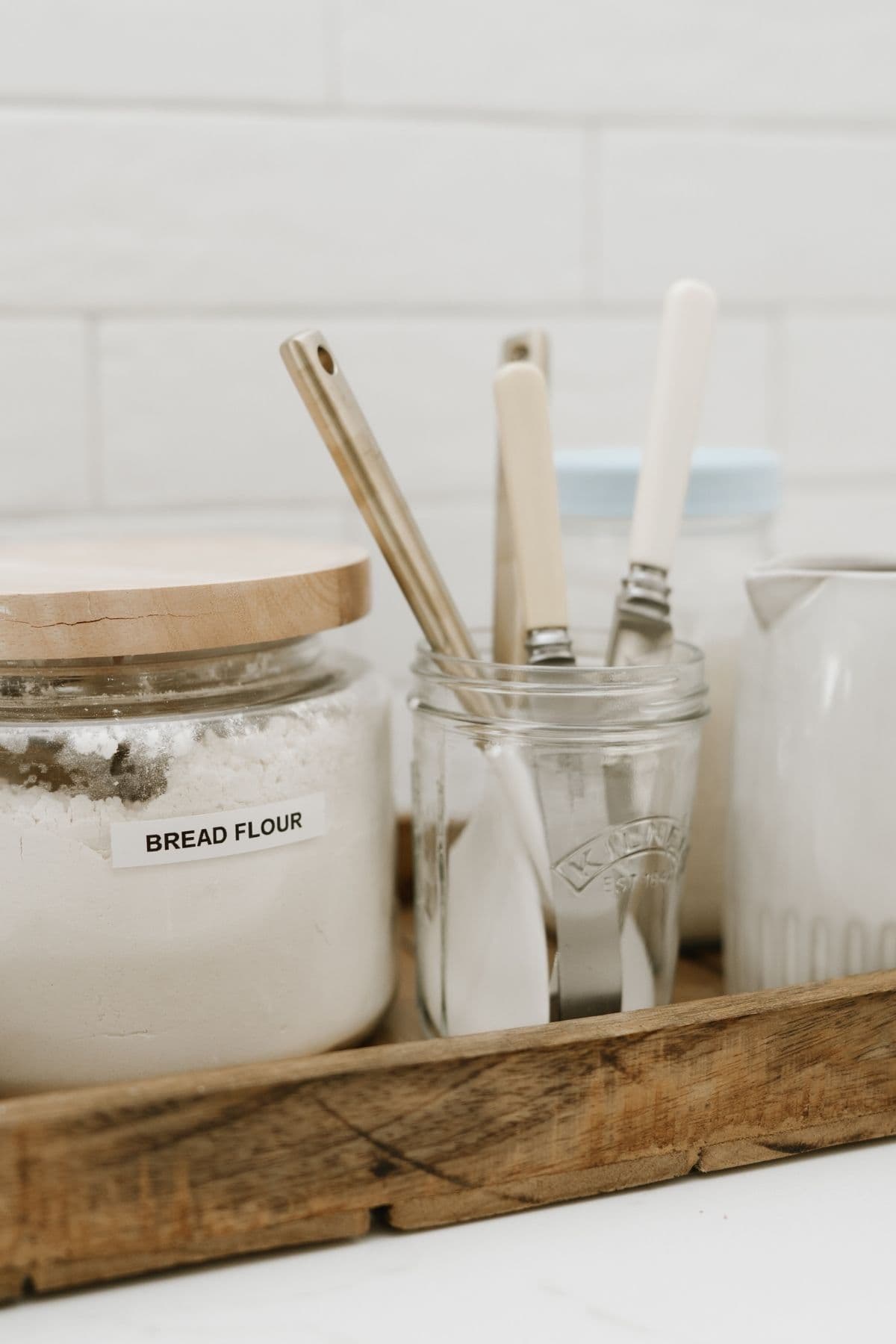 A photo showing a wooden tray containing a glass canister of bread flour with a wooden lid, a jar filled with knives to stir sourdough starter.