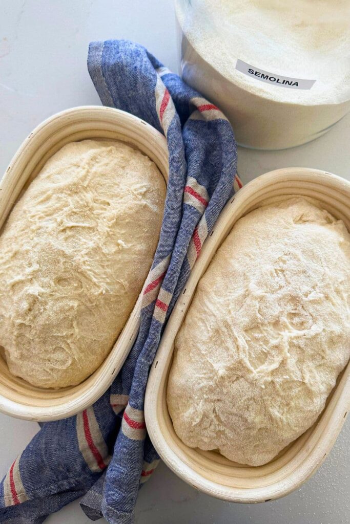 Two loaves of sourdough bread sitting in banneton baskets. There is a blue and red striped dish towel sitting between the two baskets of dough and a jar of semolina flour sitting at the back of the baskets.