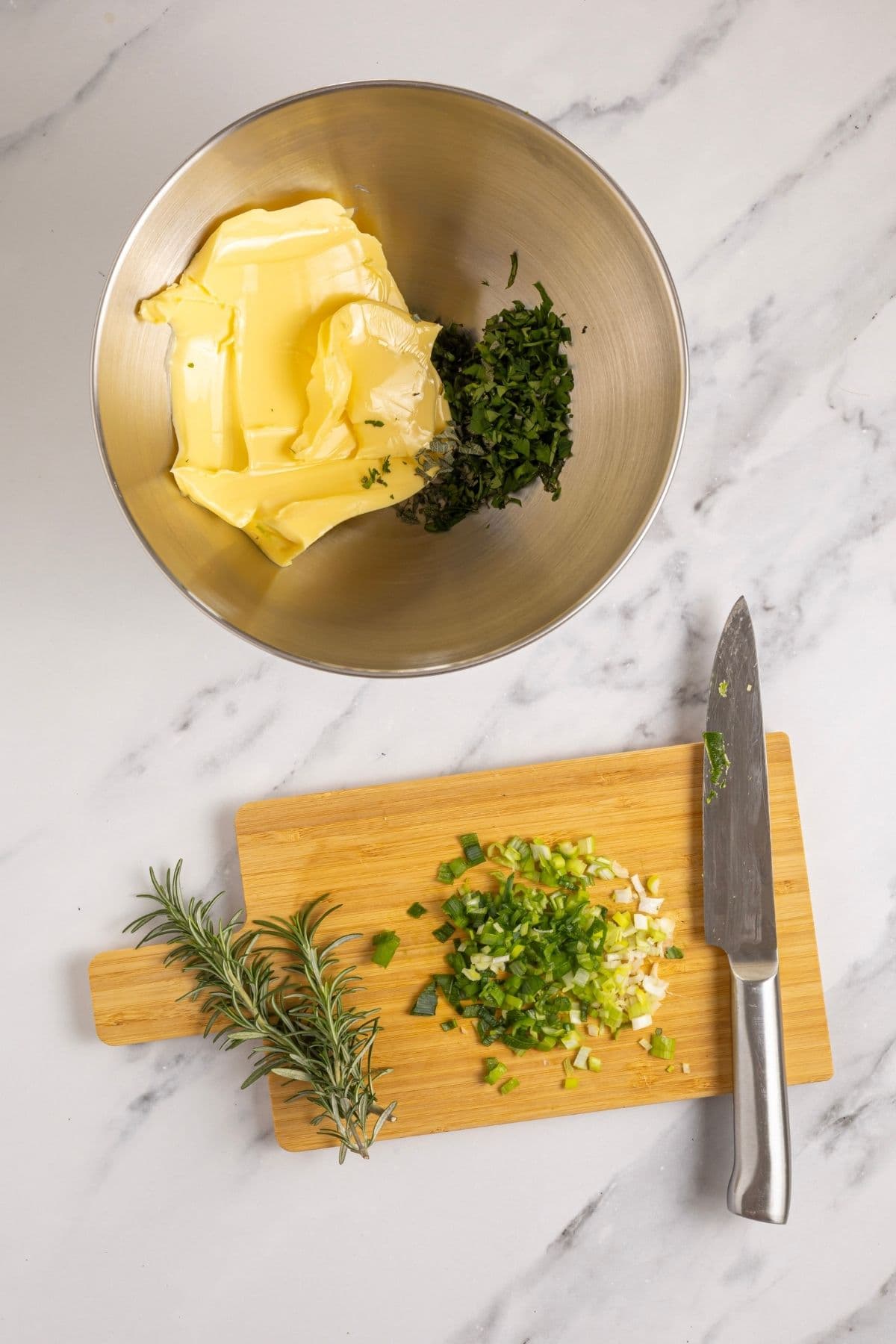 A stainless steel bowl filled with butter and finely chopped herbs. There is a also a wooden chopping board showing the herbs being chopped.