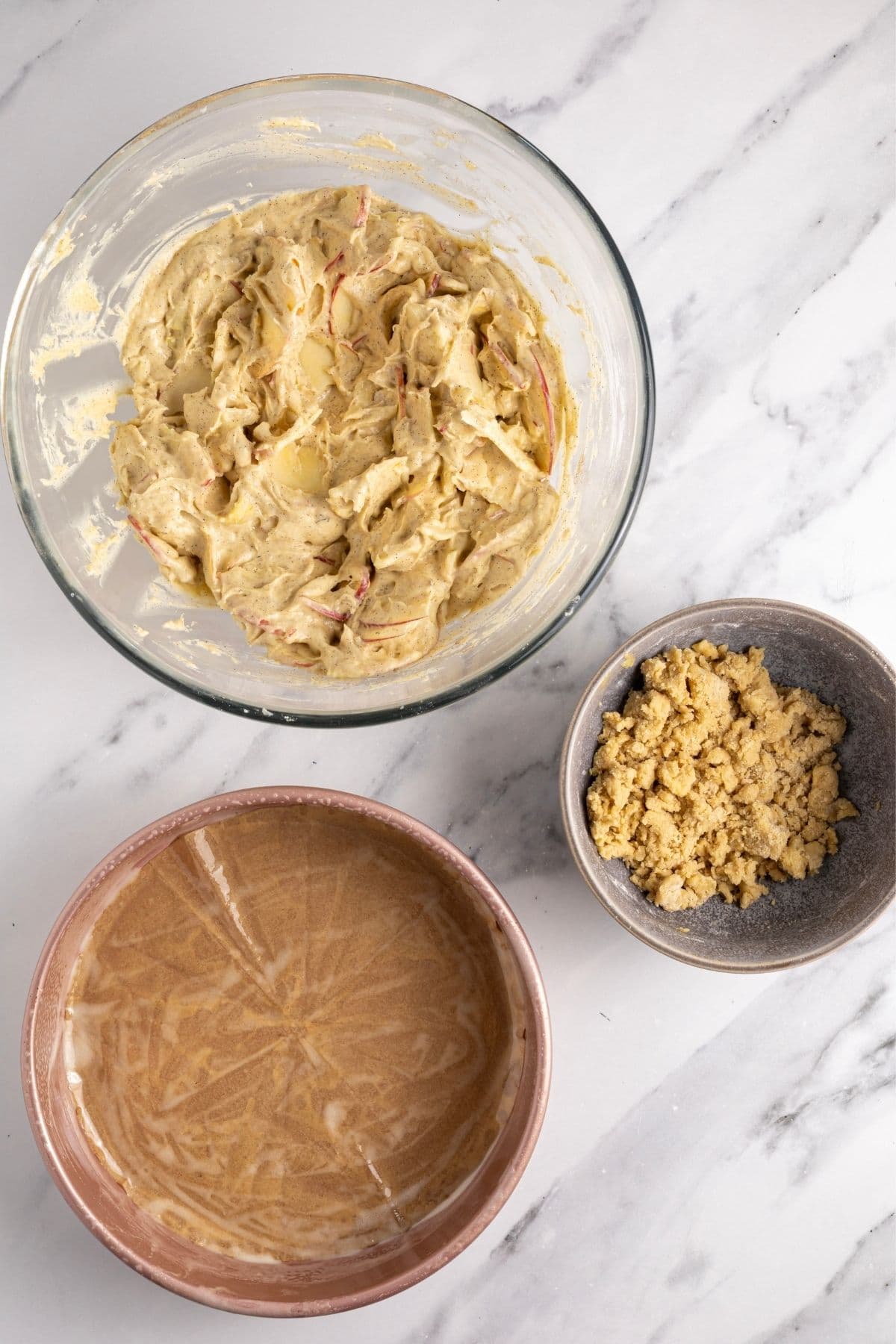 An image showing the sliced apples tossed through the cake batter, along with a small grey bowl of streusel and a round cake pan lined with parchment paper. 