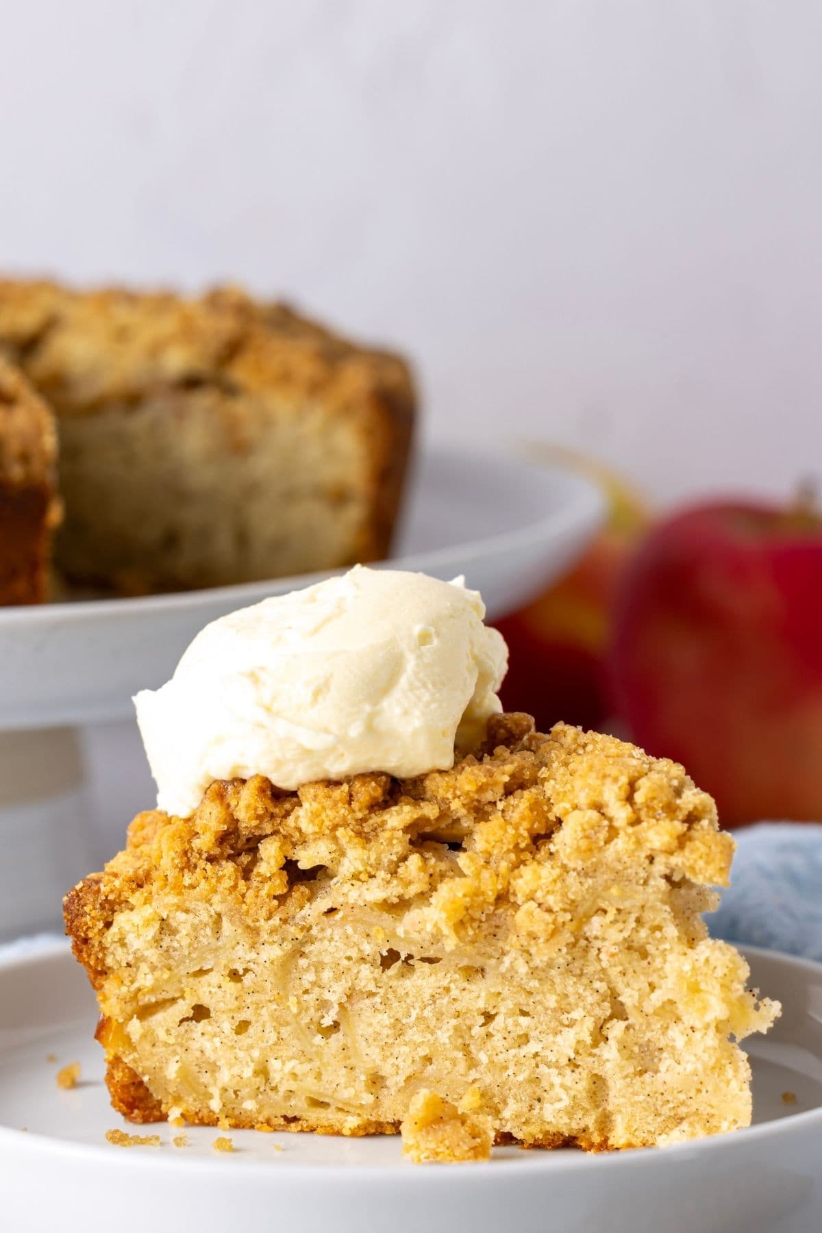 A single slice of sourdough apple cake placed on a white plate and topped with a dollop of whipped cream. There is a red apple in the background.