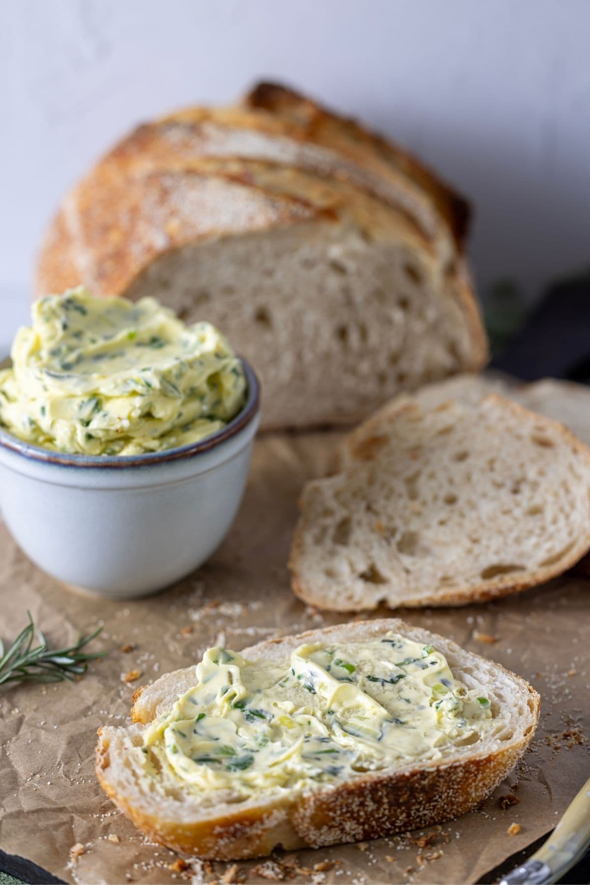 A slice of fresh sourdough bread spread with whipped herb butter. You can see the rest of the loaf in the background of the photo along with a dish of the butter.