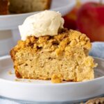 A single slice of sourdough apple cake placed on a white plate and topped with a dollop of whipped cream.