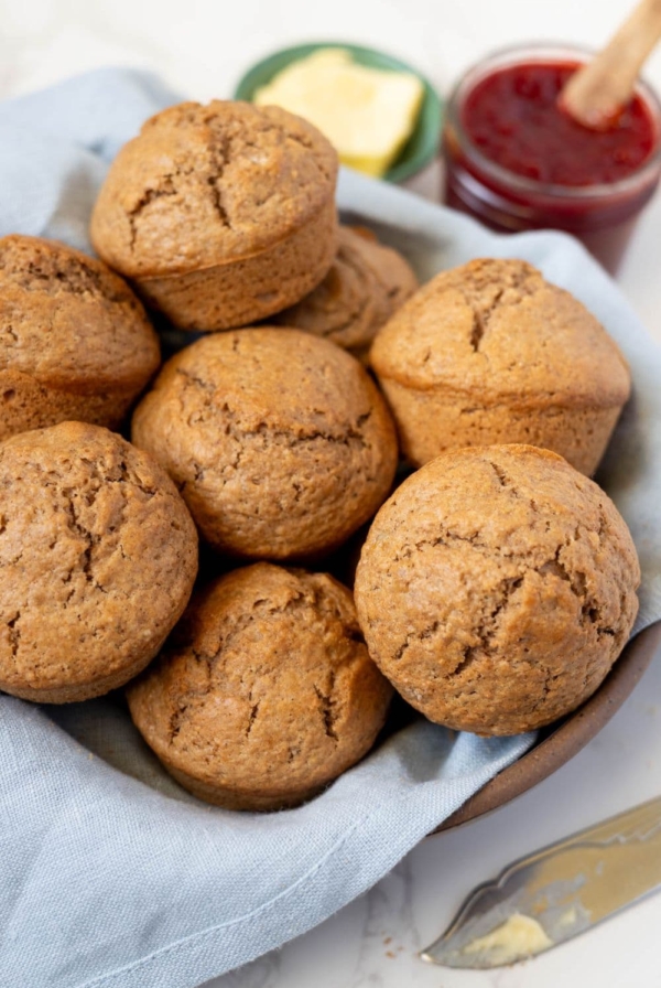 Sourdough cinnamon muffins stacked into a basket lined with a blue dish towel. There is a small dish of butter and jar of strawberry jam in the background.