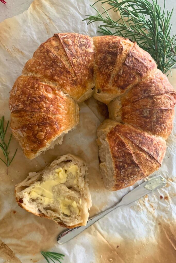 Sourdough Parmesan Bread with Rosemary baked into a round wreath shape. There is a slice cut out of the loaf that has been buttered.