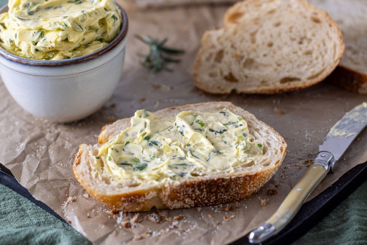 Whipped herb butter spread on a slice of fresh sourdough bread. There is a small pottery dish of herb butter in the background of the photo.