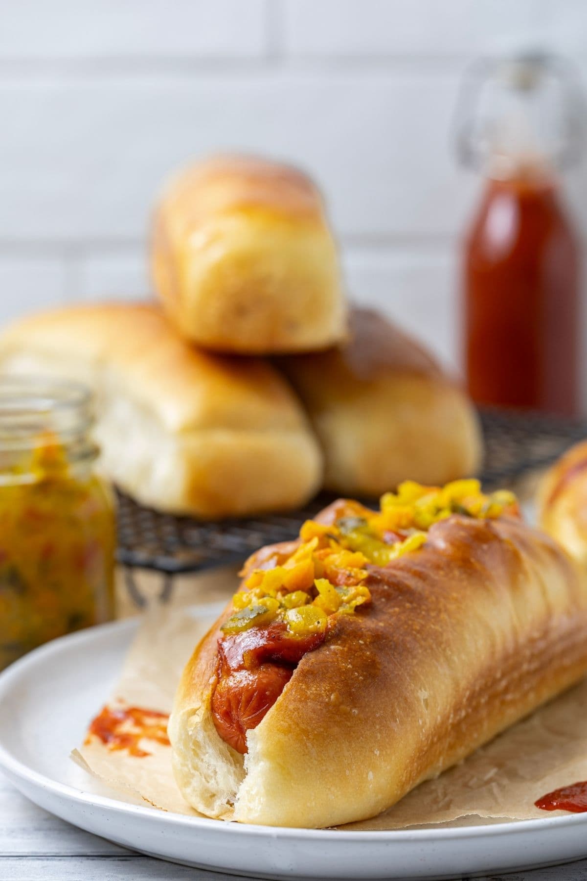 Close up photo of a sourdough hot dog bun filled with a frankfurt and topped with mustard relish. There are 3 other hot dog buns in the background of the photo and a bottle of homemade tomato ketchup.