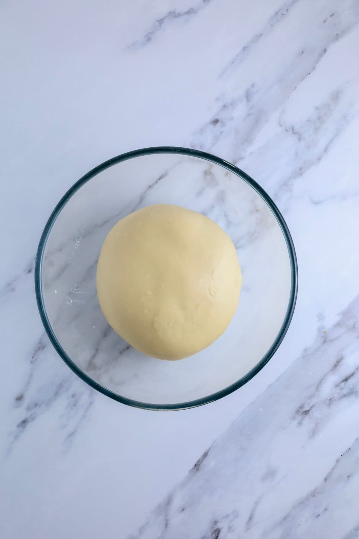 A glass bowl containing smooth sourdough hot dog bun dough that has risen during bulk fermentation and is now ready to be shaped.