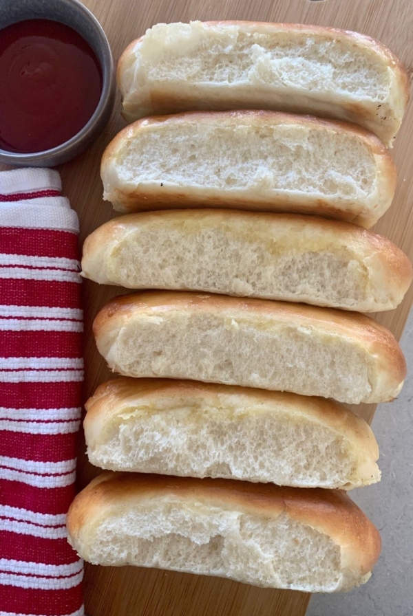 6 sourdough hot dog buns laying on their side so you can see their soft and squishy insides. There is a red striped dish towel next to them as well as a bowl of ketchup.