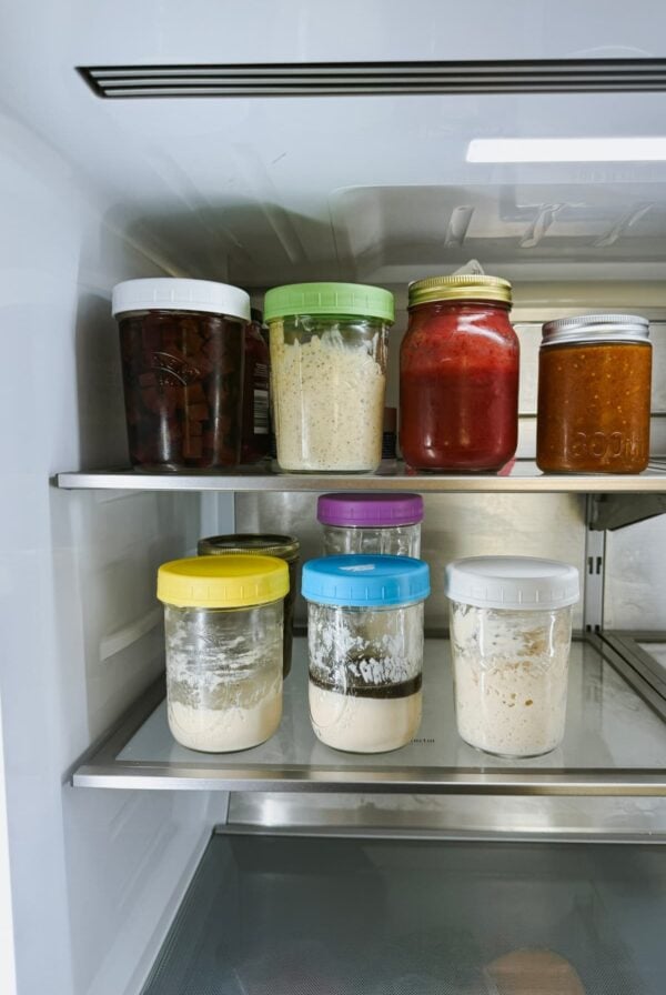 A photo showing the inside of a fridge. On the bottom shelf there are 3 jars of sourdough discard at various ages and stages. The top shelf shows an assortment of pickles and chutneys.