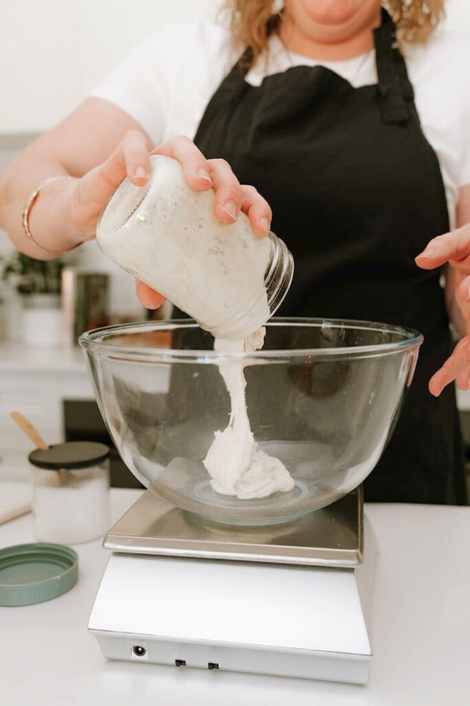 A woman wearing a black apron pouring a jar of sourdough starter into a glass bowl that is sitting on a digital scale.