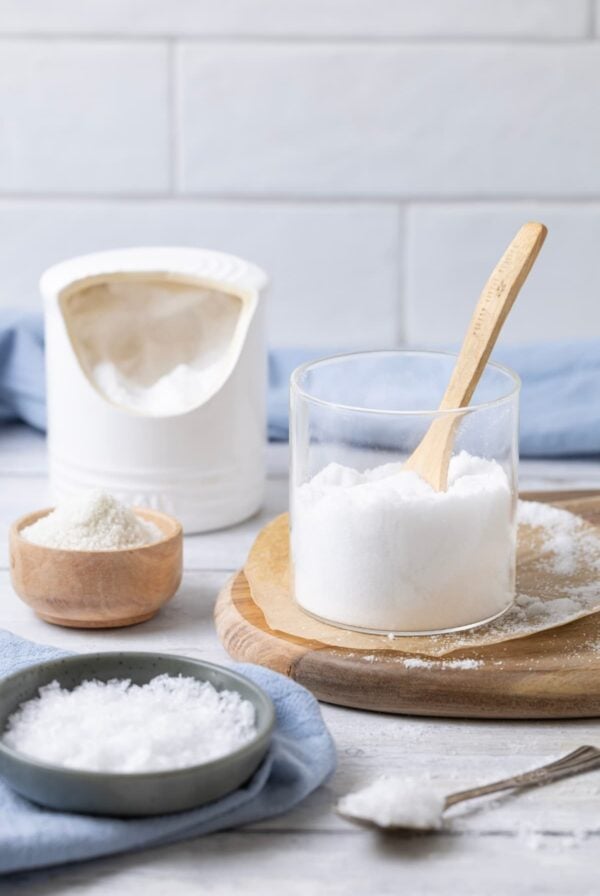 This image shows 3 different types of salt. There is a small blue bowl filled with flaky sea salt, a glass container with fine sea salt and a white salt pig at the back.