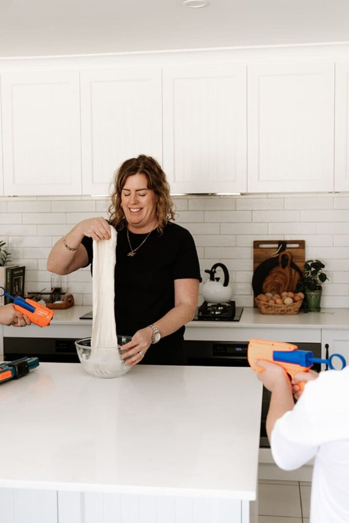 A woman in the kitchen stretching sourdough from a glass bowl on the counter.