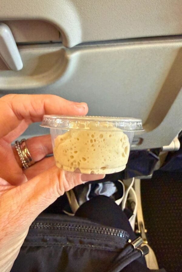 A hand holding a 2 oz container of sourdough starter in front of an aeroplane tray table.