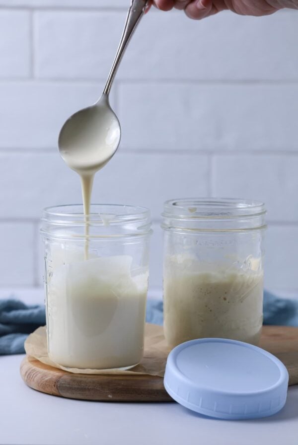 A spoon taking some runny sourdough starter out of a jar showing the liquid texture. There is a second jar next to this one and a pale blue plastic lid sitting next to them.