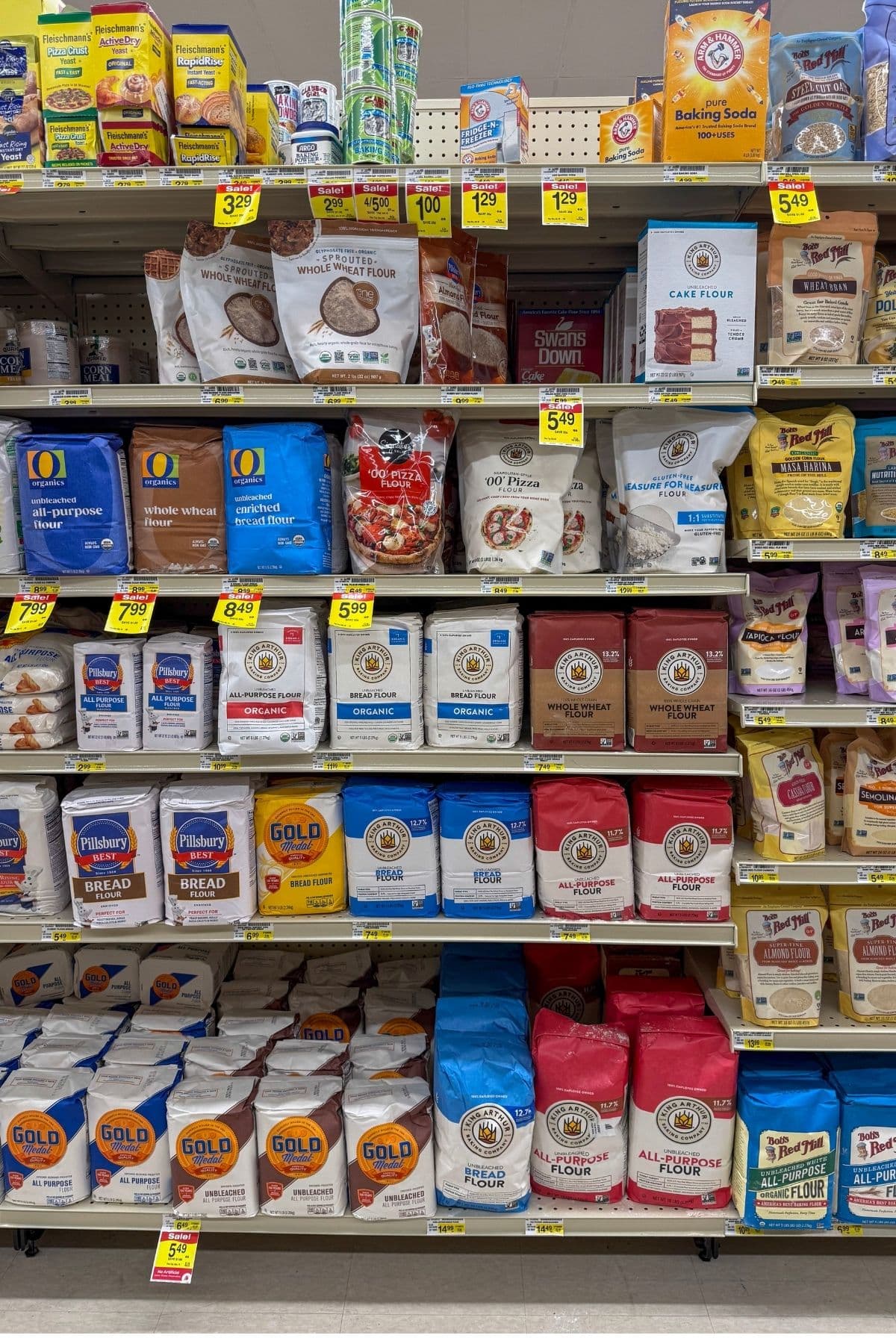 A photo of the grocery store flour aisle. The photo shows several shelves of flour including all purpose flour, bread flour, pizza flour etc. There are some yellow sale tickets displayed on some products.