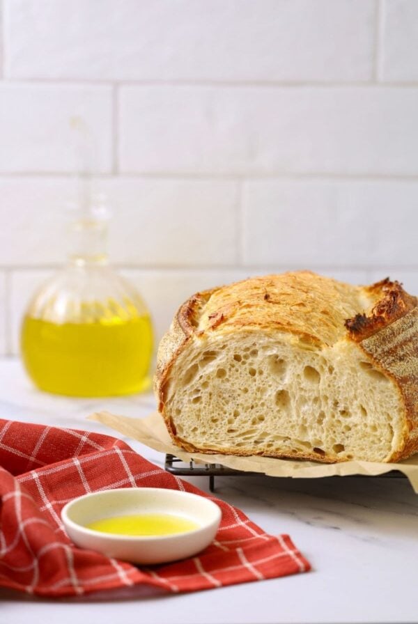 A photo showing a loaf of sourdough bread that has had olive oil added to soften the crumb and crust. There is a glass bottle of olive oil in the background, as well as a small white dish of yellow olive oil on a red checked dish towel in the foreground.