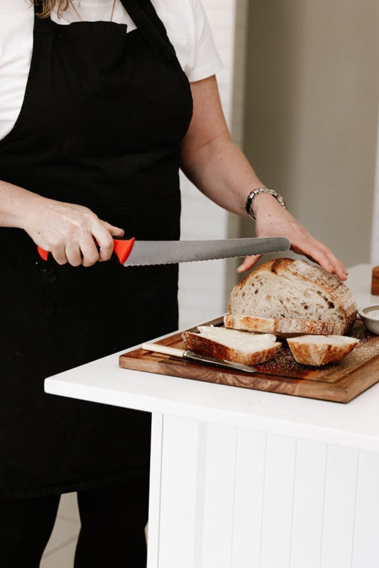 Photo showing a woman using a red handled Mercer bread knife to slice a loaf of sourdough bread.