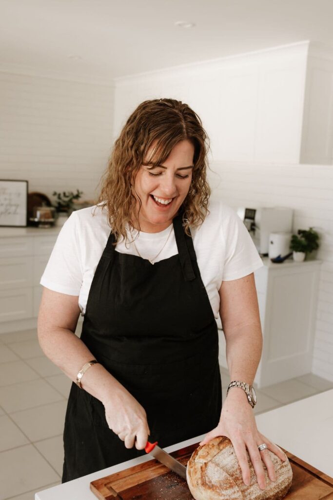 A photo of Kate from the Pantry Mama smiling and slicing a loaf of sourdough bread with a red handled Mercer bread knife.