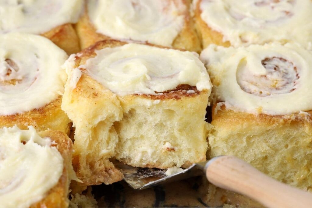 A close up photo of sourdough cinnamon rolls topped with vanilla cream cheese frosting. One of the rolls is being lifted out of the tray by a wooden handled cake slice.