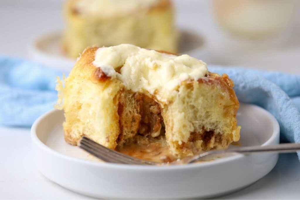 A sourdough cinnamon roll sitting on a white plate. The cinnamon roll is filled with gooey cinnamon brown sugar filling and topped with vanilla cream cheese filling.