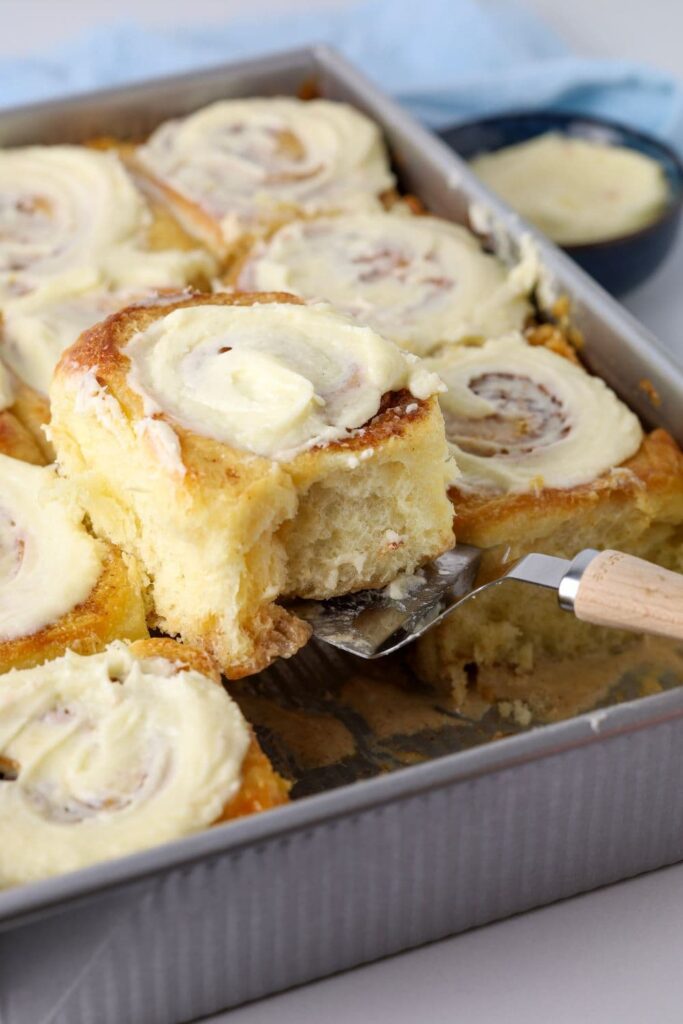 A tray of sourdough cinnamon rolls topped with vanilla cream cheese frosting. One of the cinnamon rolls is being lifted out of the baking pan.