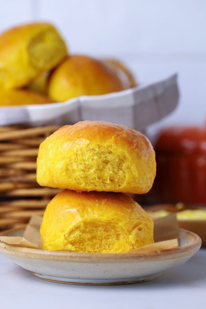 2 sourdough pumpkin dinner rolls stacked on top of each other on a small plate in front of a wicker basket full of sourdough pumpkin dinner rolls.
