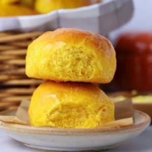 2 sourdough pumpkin dinner rolls stacked on top of each other on a small plate in front of a wicker basket full of sourdough pumpkin dinner rolls.