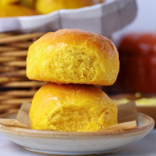 2 sourdough pumpkin dinner rolls stacked on top of each other on a small plate in front of a wicker basket full of sourdough pumpkin dinner rolls.