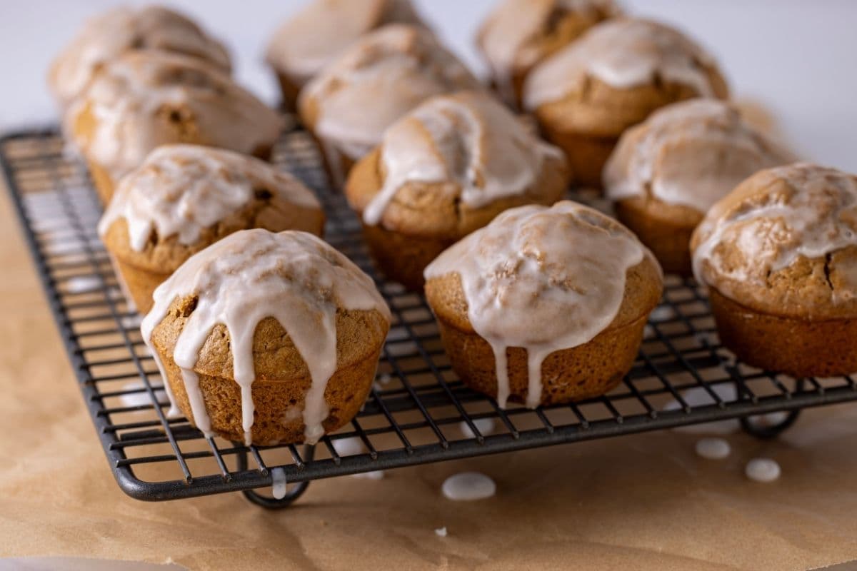 A batch of 12 sourdough gingerbread muffins sitting on a wire cooling rack. The muffins have been drizzled with a snowy white glaze that is dripping down onto the counter underneath.