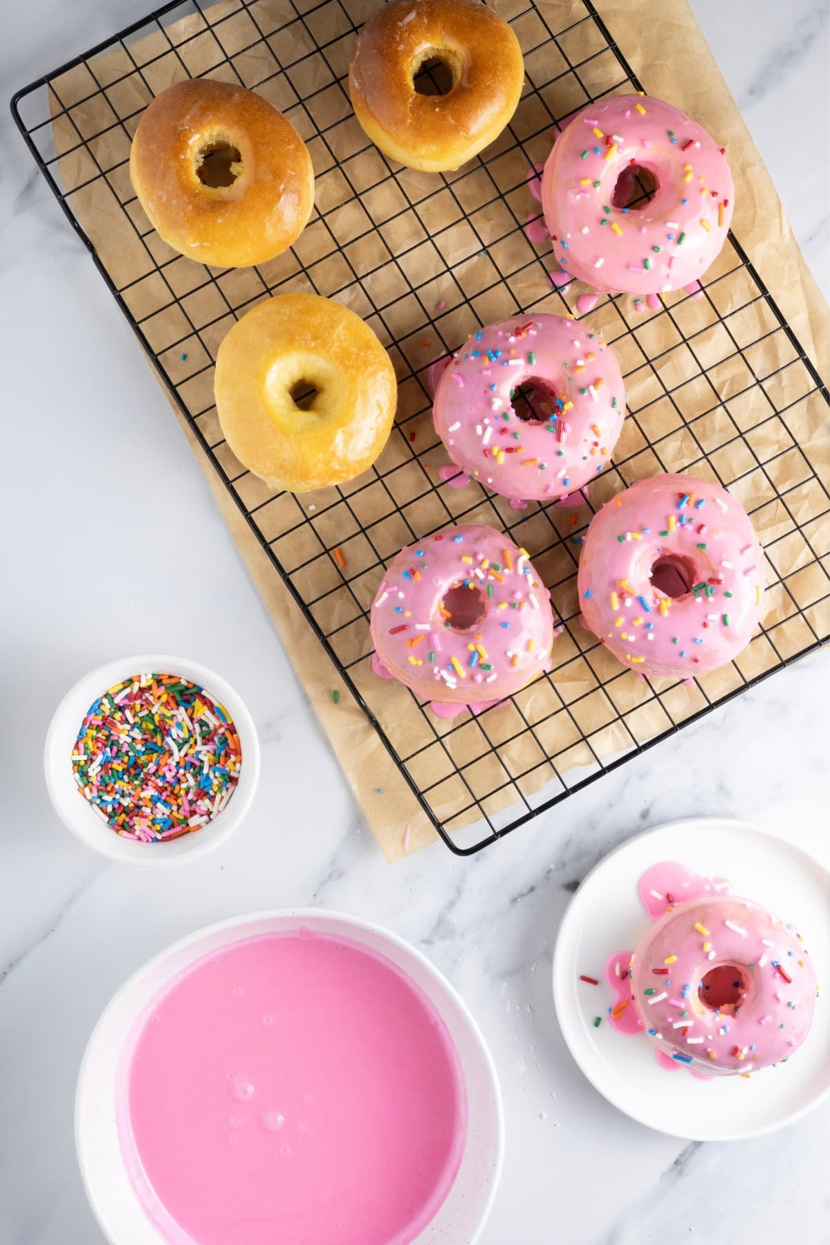 A flat lay image showing some freshly baked sourdough donuts sitting on a wire cooling rack. There is a bowl of bright pink milk glaze and a small bowl of rainbow sprinkles. Some of the donuts have been glazed whereas others are still waiting.