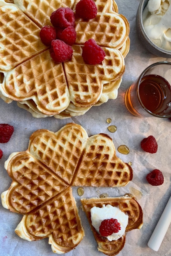 A flat lay of sourdough discard waffles made in a round waffle maker. The waffles are displayed on a board with fresh raspberries, whipped cream and maple syrup.