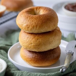 A stack of 3 golden brown sourdough bagels sitting on a white plate.