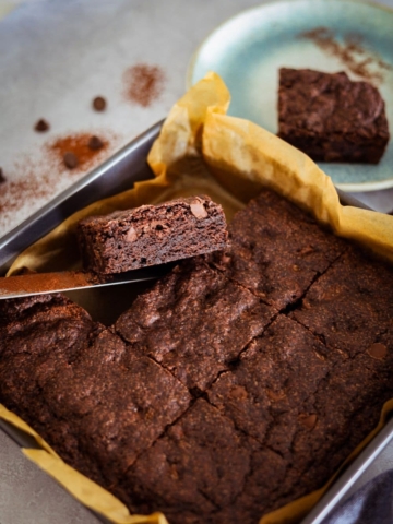 A tray of rich, fudgy chocolate sourdough brownies. They have been sliced into squares in the tin and there is a piece of brownie lifted up so you can see the fudgy crumb inside.