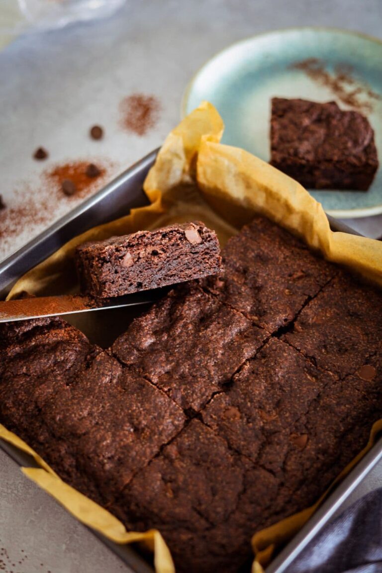 A tray of rich, fudgy chocolate sourdough brownies. They have been sliced into squares in the tin and there is a piece of brownie lifted up so you can see the fudgy crumb inside.