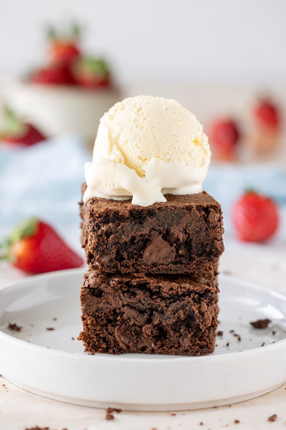 A vertical photo showing two sourdough brownies stacked on top of one another and topped with a scoop of vanilla ice cream. You can see ripe red strawberries in the background of the image.