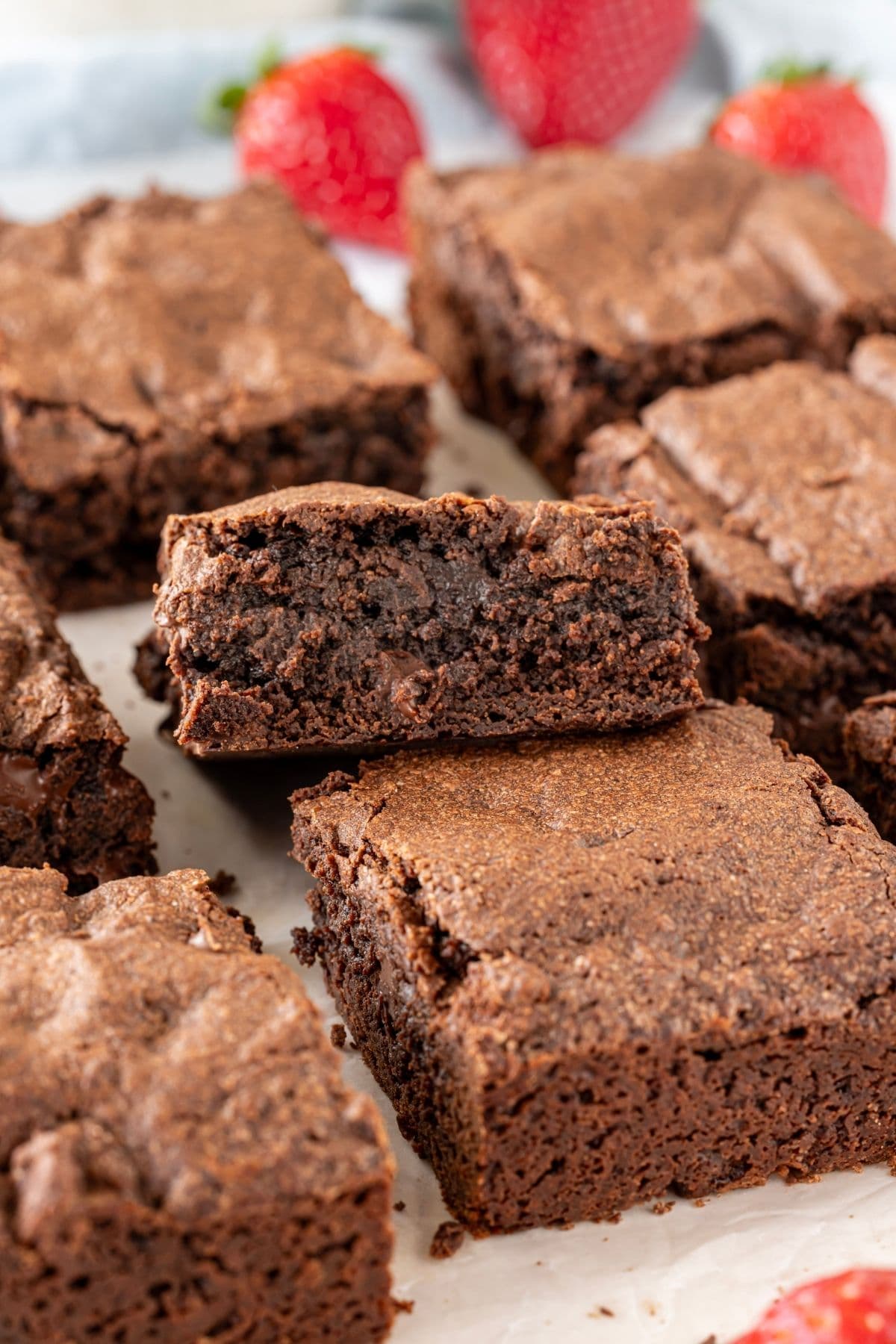 A vertical image showing a close up of a square of sourdough brownie. You can see the rich, fudgy center and the crackly, meringue tops. There are ripe, red strawberries featured in the background of the image.