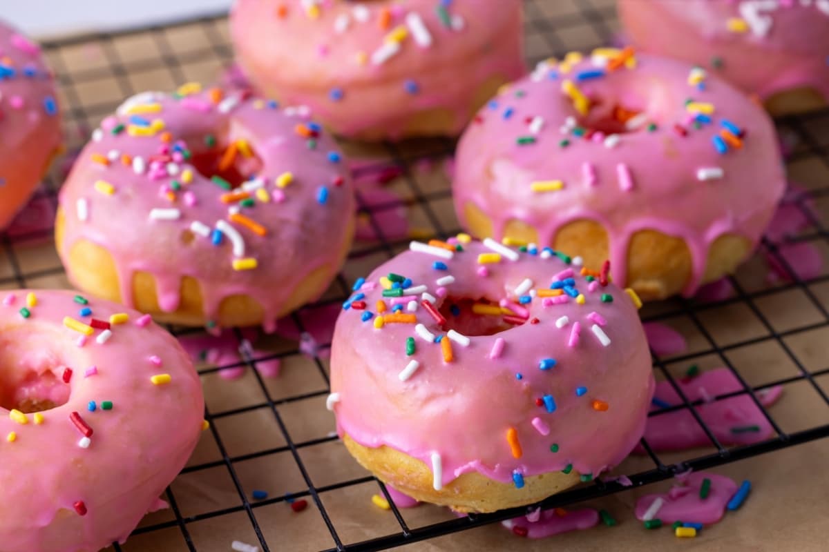 A black wire cooling rack topped with 7 baked sourdough donuts drizzled in bright pink glaze and topped with rainbow sprinkles. 