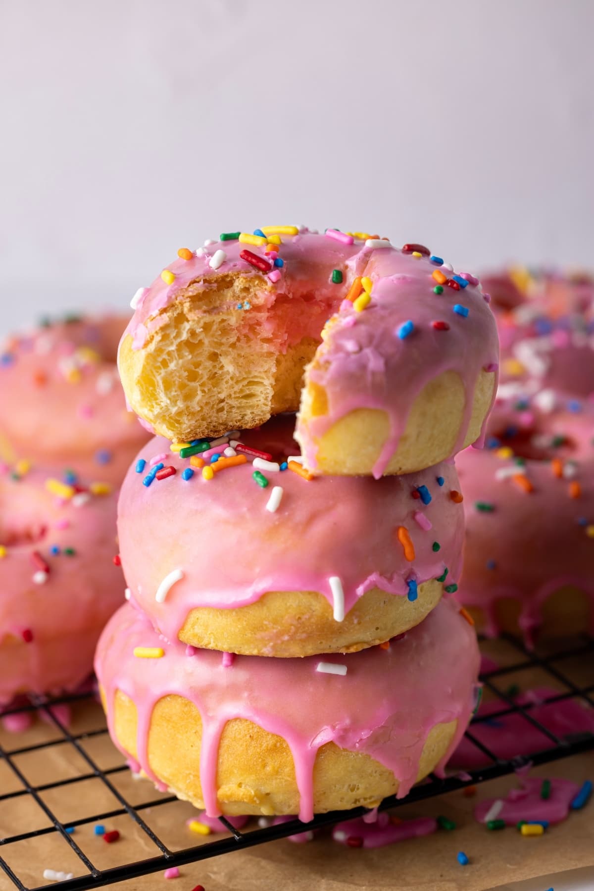 A stack of 3 baked sourdough donuts drizzled in pink glaze. You can also see other donuts in the background of the stack. They are all sitting on a black wire cooling rack.