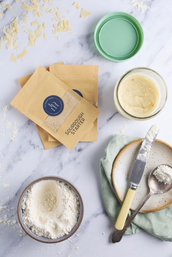 A flat lay image showing small packets of purchased sourdough starter laying next to a jar of homemade sourdough starter. There is also a small knife used to stir the starter and spoon with flour on it showing the process of making a sourdough starter.