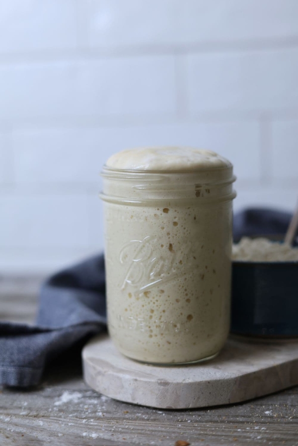 A jar of sourdough starter sitting on a small marble board surrounded by a blue dish towel. The sourdough starer is at peak and about to overflow over the top of the jar.