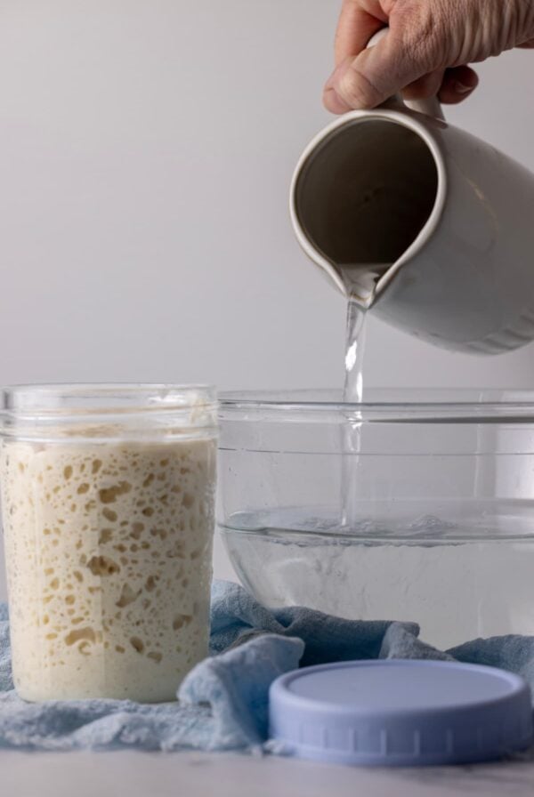 A bubbly jar of sourdough starter sitting next to a glass bowl of water. There is a jug of water being poured into the bowl from above.