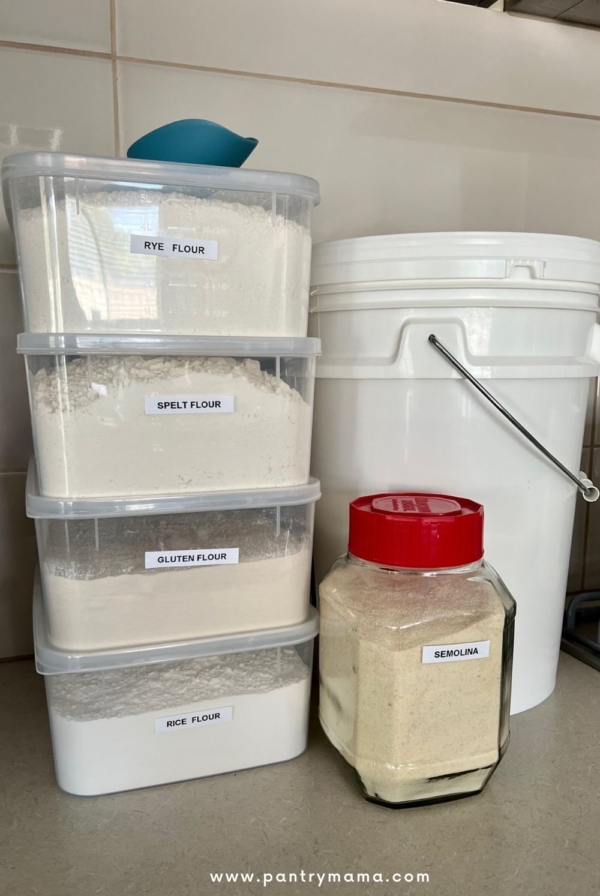 4 plastic containers with labelled with different types of flour stacked on top of one another. Next to them is a food safe bucket full of flour and a glass jar with a red lid containing semolina flour.
