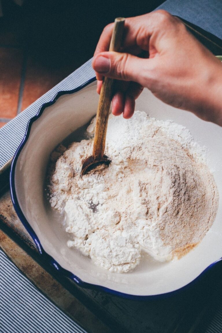 Wooden chopping board with a blue rimmed bowl sitting on top. The bowl is filled with flour and water and being stirred with a wooden spoon.